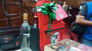 Una mochila roja con el logo del PRI y un moño verde contiene una botella de alcohol. Se encuentra sobre una mesa junto a otra botella de licor.