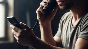 Primer plano enfocado en una mano sosteniendo un celular, con la silueta borrosa de una persona con capucha oscura al fondo.