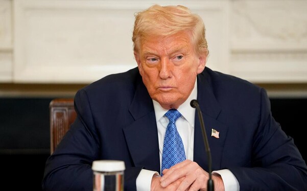 Donald Trump emitiendo un mensaje portando un saco azul y una corbata azul. Sentado en una silla café con un fondo blanco detrás.