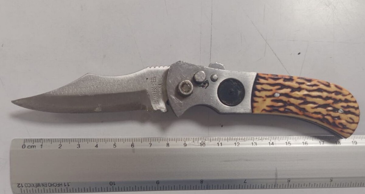 Navaja de veinte centímetros con mango de madera y una regla metálica debajo, ambas sobre una mesa blanca