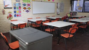 Se observan unos pupitres  y un escritorio, dentro de un salón de clases.
