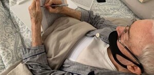 Se observa a un hombre canoso con un cubrebocas negro, firmando unos papeles.