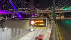 Letrero electrónico nocturno sobre un túnel indica '5 CARRILES DE SUR A NORTE'. De fondo destaca el puente Matute Remus con luces púrpuras.