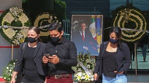 Personas de negro y con cubrebocas caminan frente a un retrato de Aristóteles Sandoval rodeado de coronas florales en lo que parece un memorial.
