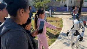 Una niña con conjunto rosa interactúa con un robot humanoide blanco en un parque, mientras una mujer graba la escena con su teléfono celular.