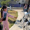 Una niña con conjunto rosa interactúa con un robot humanoide blanco en un parque, mientras una mujer graba la escena con su teléfono celular.