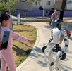 Una niña con conjunto rosa interactúa con un robot humanoide blanco en un parque, mientras una mujer graba la escena con su teléfono celular.