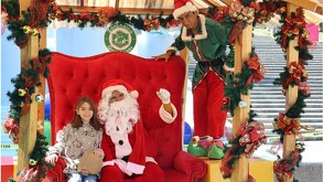 Santa Claus sentado en un gran trono rojo con una niña y un duende en un set navideño decorado con guirnaldas del Zoológico Guadalajara.