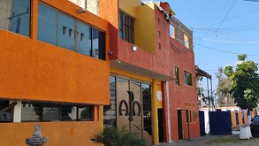 Fachada de un edificio de dos niveles pintado en colores naranja y amarillo vibrantes, con grandes ventanales y un portón metálico con símbolos.