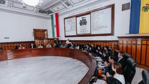 Una sesión de cabildo en una sala solemne con una mesa circular de madera. Al fondo, un muro con nombres ilustres.