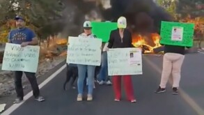 Cuatro personas sostienen carteles de protesta en una carretera bloqueada por fuego y humo; los mensajes exigen justicia y la aparición de personas.