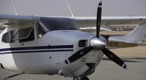 Vista de frente de una aeronave blanca con rayas azules tipo Cessna C172 con hélices negras estacionada en una pista de aterrizaje