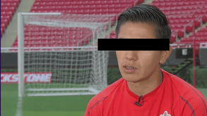Primer plano de un hombre joven con camiseta deportiva roja en un estadio de fútbol; sus ojos están cubiertos por una barra negra de censura.