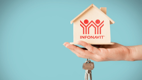 Una mano sostiene una pequeña casa de madera con el logotipo de INFONAVIT impreso en rojo, junto a un juego de llaves, sobre un fondo azul claro.