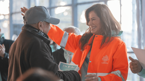 Una funcionaria con chaqueta naranja del Gobierno de Guadalajara sonríe mientras saluda a un ciudadano de la tercera edad en un evento público.