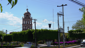 Una torre de campanario de ladrillo con un reloj resalta sobre el follaje verde de una plaza pública, bajo un cielo azul con cables de luz al frente.
