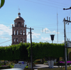 Una torre de campanario de ladrillo con un reloj resalta sobre el follaje verde de una plaza pública, bajo un cielo azul con cables de luz al frente.