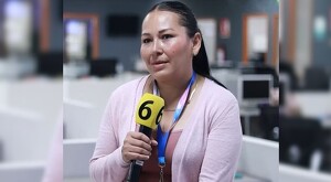 Beatriz Uscanga viendo a un costado mientras sostiene un micrófono de Canal 6 con el fondo desenfocado de las oficinas de Telediario Guadalajara