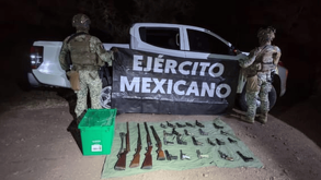 Dos soldados del Ejército Mexicano custodian un arsenal de armas largas y cortas decomisadas, desplegadas en el suelo junto a una camioneta blanca.