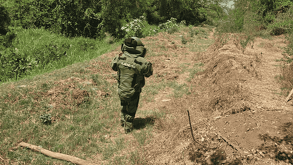 Un elemento del Ejército Mexicano, equipado con un traje especial antibombas de color verde, camina por un terreno baldío con vegetación.