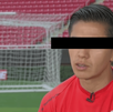 Primer plano de un hombre joven con camiseta deportiva roja en un estadio de fútbol; sus ojos están cubiertos por una barra negra de censura.