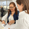 Una mujer con chaleco azul y lentes revisa documentos junto a otra persona en una oficina de atención ciudadana con ventanillas al fondo