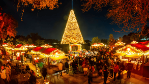 Un gran árbol de Navidad iluminado domina una plaza nocturna llena de gente y puestos de madera decorados con luces doradas en un mercado festivo.