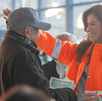 Una funcionaria con chaqueta naranja del Gobierno de Guadalajara sonríe mientras saluda a un ciudadano de la tercera edad en un evento público.
