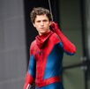 El actor Tom Holland posa como Spider-Man con el traje rojo y azul, pero sin la máscara puesta. Sostiene un hilo de telaraña con su mano derecha.