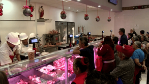 Carniceros con cascos blancos atienden a una fila de clientes tras un mostrador con cortes de carne roja, en un local decorado con esferas navideñas.