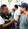 Pablo Lemus Navarro estrecha la mano de un hombre con camiseta de tirantes frente a un muro blanco. Hay personas y militares en el fondo.