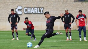 Jugadores del equipo Atlas FC entrenan en una cancha de césped; un futbolista patea el balón mientras otros observan al fondo frente a un muro.