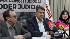 Un hombre habla frente a micrófonos de prensa en una mesa; detrás hay una lona sobre el Poder Judicial y una bandera blanca con el logo de Morena.