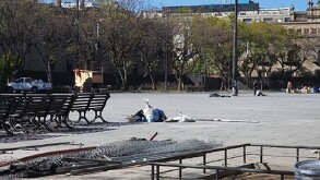 Una amplia plaza pública de concreto en el centro histórico, con hileras de bancas de madera a la izquierda y material de construcción en primer plano.