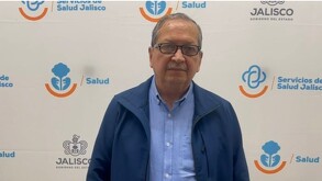 Un hombre con anteojos y chaqueta azul posa frente a un muro blanco con los logotipos oficiales de 