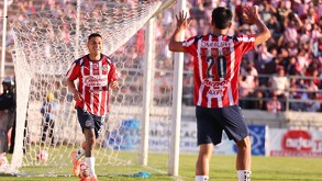 Un jugador del Club Guadalajara con el uniforme rojiblanco corre celebrando un gol frente a la portería, con la afición de fondo en el estadio.