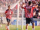 Un jugador del Club Guadalajara con el uniforme rojiblanco corre celebrando un gol frente a la portería, con la afición de fondo en el estadio.