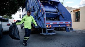 Se observa un hombre vestido con camisa verde con una boolsa negra y atrás un camión de basura.