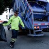 Se observa un hombre vestido con camisa verde con una boolsa negra y atrás un camión de basura.