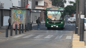 Un autobús verde de la ruta 52 se aproxima a un cruce peatonal en una calle de la ciudad, junto a un puesto callejero decorado con un mural artístico.
