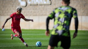 Un jugador de fútbol del equipo Atlas con uniforme rojo entrena en una cancha de pasto, preparándose para patear el balón durante el día.