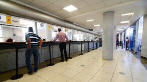 Varias personas esperan frente a las ventanillas de una oficina recaudadora gubernamental, con cajas numeradas y separadores de fila negros.