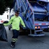 Se observa un hombre vestido con camisa verde con una boolsa negra y atrás un camión de basura.