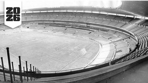Una fotografía en blanco y negro de las gradas vacías de un gran estadio de fútbol, con un logotipo en la esquina que indica el año 