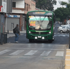 Un autobús verde de la ruta 52 se aproxima a un cruce peatonal en una calle de la ciudad, junto a un puesto callejero decorado con un mural artístico.