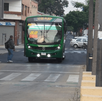 Un autobús verde de la ruta 52 se aproxima a un cruce peatonal en una calle de la ciudad, junto a un puesto callejero decorado con un mural artístico.
