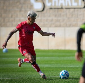 Un jugador de fútbol del equipo Atlas con uniforme rojo entrena en una cancha de pasto, preparándose para patear el balón durante el día.