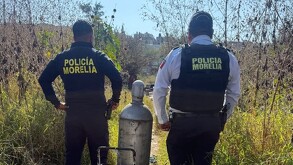 Dos agentes de la Policía de Morelia aparecen de espaldas en un terreno baldío junto a un tanque de gas metálico que se encuentra en el suelo.