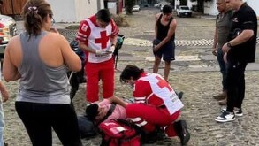 Paramédicos con uniformes rojos de la Cruz Roja brindan primeros auxilios a una persona tendida en el suelo de una calle empedrada ante testigos.