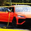 Un auto deportivo naranja con impactos de bala en el parabrisas está acordonado; un círculo superpuesto muestra boletos de rifa de colores.